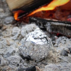 Apple wrapped tightly in foil sitting in hot campfire coals away from flames.