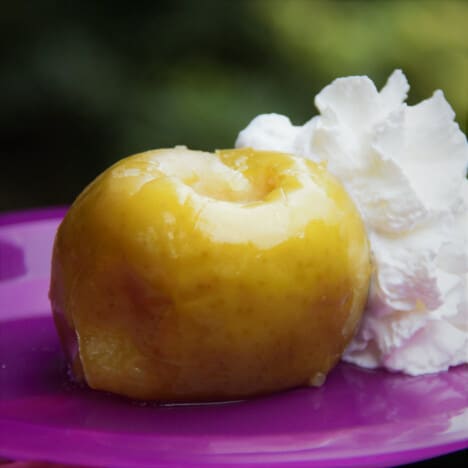 Yellow baked apple resting on a bright purple plate next to a fluffy, white mound of whipped cream.