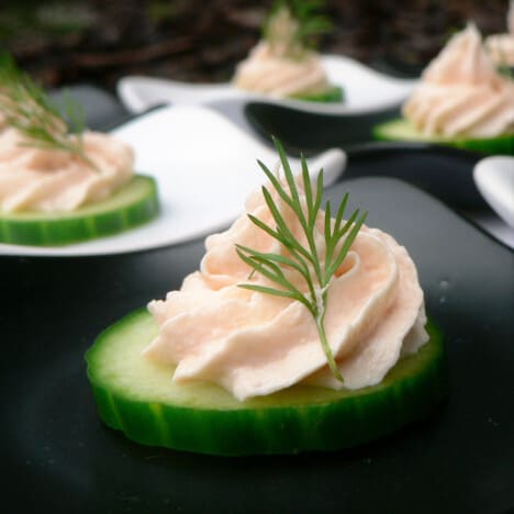 Close up of pale pink smoked salmon mousse swirl piped on a cucumber slice and garnished with sprig of green dill.