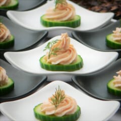 Pale pink swirls of smoked salmon mousse piped onto single cucumber slices, each garnished with a green sprig of dill and served on an individual plate.