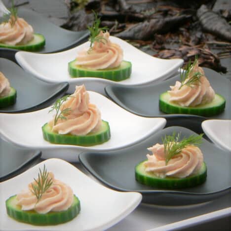 Swirls of pale pink smoked salmon mousse piped onto single cucumber slices garnished with sprigs of green dill and offered on individual plates as finger food.