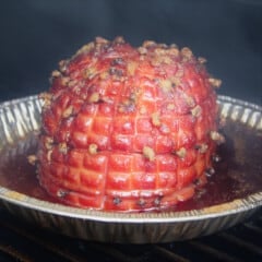 Glazed ham studded with cloves sitting in apple cider in a pie pan on a grill, surrounded by smoke.