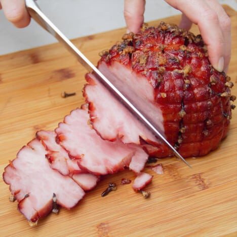 Finished glazed ham studded with cloves resting on a cutting board as slices are carved.