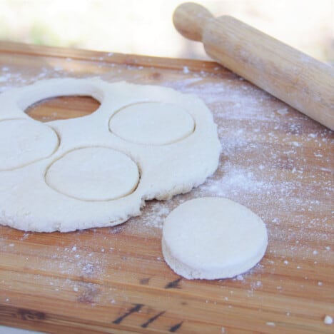 Flat sheet of dough on a wooden cutting board with round scones cut out in preparation to cook.