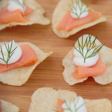 Close up of a potato chip topped by a small slice of smoked salmon, a piped dollop of cream cheese and a tiny sprig of dill.