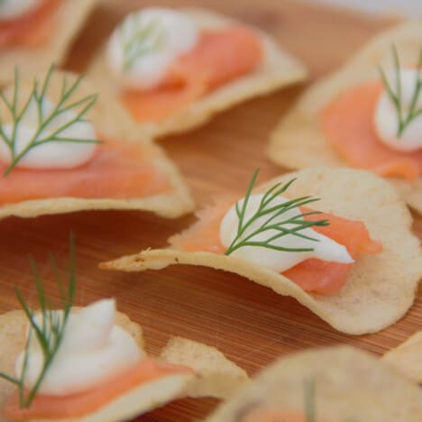 Close up of a potato chip topped by a small slice of smoked salmon, a piped dollop of cream cheese, and a tiny sprig of dill.