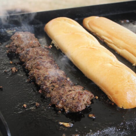 One single long burger patty sitting next to the two halves of the bread roll toasting on the flat top grill.