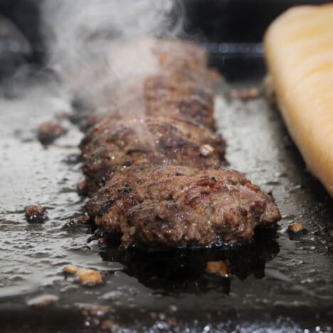 A long burger patty cooking on a flat top grill with steam coming off of it.