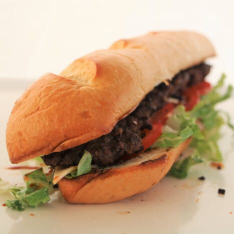 A fully completed foot long burger sitting on a white chopping board with the lettuce spilling out.