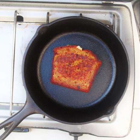 Overhead view of a slice of banana bread frying in a cast iron skillet over a gas camp stove.