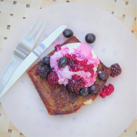 Overhead view of a slice of fried banana bread topped with yogurt and mixed berries on a white plate next to a fork and knife.