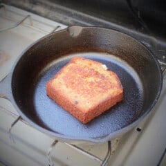 A slice of banana bread fries in a cast iron skillet on a gas camp stove.