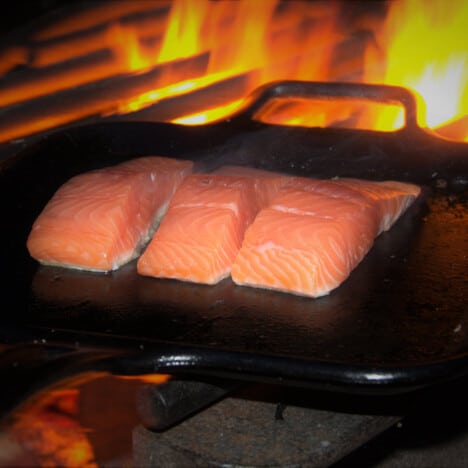Three salmon slices frying on a square, cast iron skillet sitting on a grill over a fire.