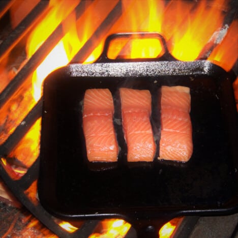 Three individual portions of salmon cooking on a cast iron skillet resting on a grill over a fire.