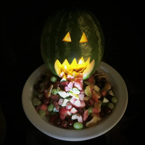 Watermelon jack-o-lantern in the dark, light from inside it is shining on fruit spilling out its mouth and down a platter.