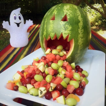 Carved watermelon jack-o-lantern resting upright on a large plate with fruit spilling out its mouth and down the plate.