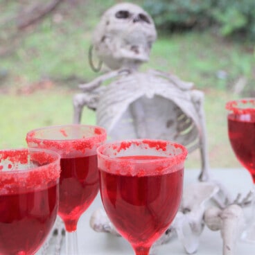 Wine glasses of red jello with a red rim displayed on a serving table with a plastic skeleton.