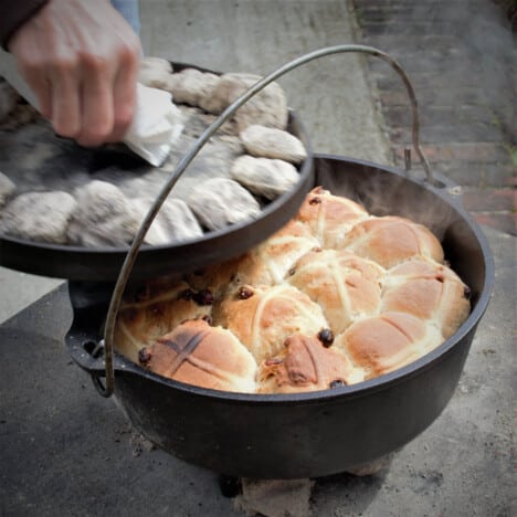 Steam coming off fully baked, golden brown hot cross buns in black Dutch oven, a person's hand removing the lid covered in coals.