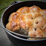 Golden brown fully baked hot cross buns with lighter crosses on each bun, nestled in a black Dutch oven with one bun removed.