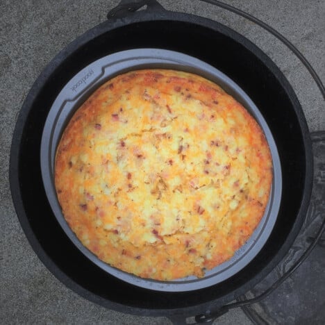 Overhead view of a fully baked, golden brown quiche in a pie pan inside an open Dutch oven with the oven lid next to it.
