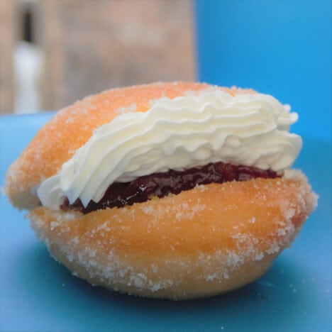 Close up of a single completed, golden brown Kitchener bun crusted with sugar and filled with piped pastry cream atop jam.
