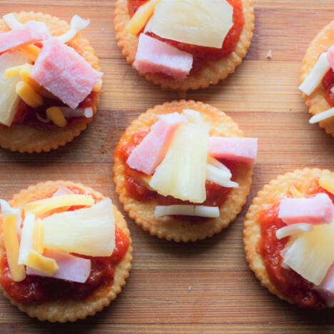 Looking down on ham and pineapple pizza crackers laid out on a chopping board.