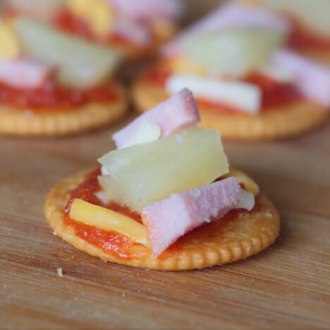 Close up of a cracker toped with pizza sauce, shredded cheese, ham, and pineapple.