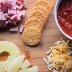 Crackers, pizza sauce, diced ham, pineapple, and shredded cheese laid out on a wooden chopping board ready to be made into mini pizzas.