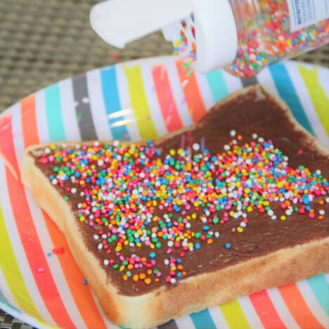 A slice of bread with Nutella spread on it and sprinkles being added.