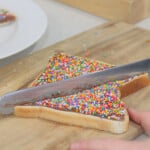 A piece of fairy bread being cut in half to make a triangle on a wooden chopping board.