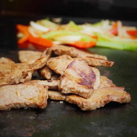 Close up of cooked marinated beef strips sitting on a griddle with chopped onions and bell peppers in the background.