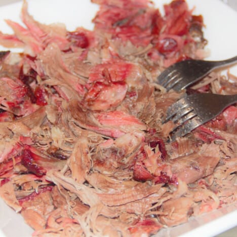 Large bowl of fully cooked and pulled pork with two silver forks used for shredding, resting on the side.
