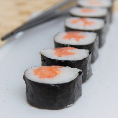 Close up of a row of sliced sushi pieces, pink smoked salmon surrounded by white sushi rice wrapped in dark green seaweed.