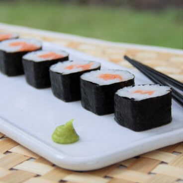 Sliced sushi pieces lined up on a white dish next to black chopsticks and a dollop of bright green wasabi.