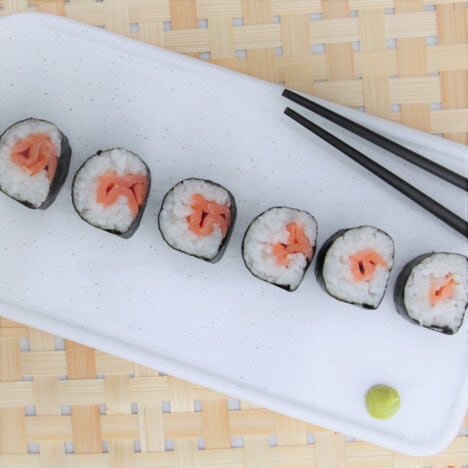 Overhead view of sliced sushi pieces lined up on a white dish next to black chopsticks and a dollop of bright green wasabi.