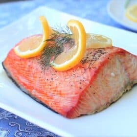 Cooked salmon filet garnished with two thin, twirled lemon slices and a dill sprig resting skin down on a white platter.