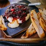 Smoked brie wheel oozing melted cheese, topped and dripping with chunky cranberry chutney, resting on a serving platter next to slices of toasted bread.