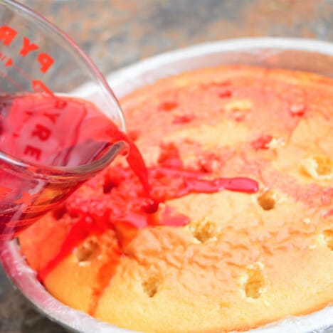 Liquid red jello being poured onto a hole covered cake in a plastic lined cake tin.