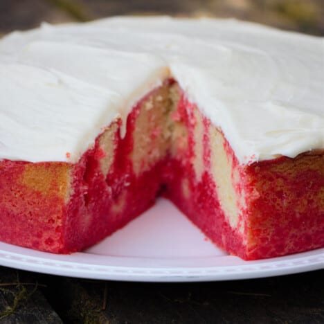 A round strawberry poke cake with a large slice cut out exposing the red jello design within.