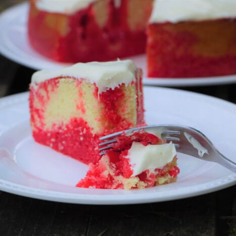 A wedge slice of strawberry poke cake with a fork having cut off the first bite, while the rest of the cake is in the background.