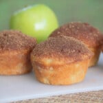 Cooked and topped apple and cinnamon muffins on a serving board with a raw apple ready to eat.