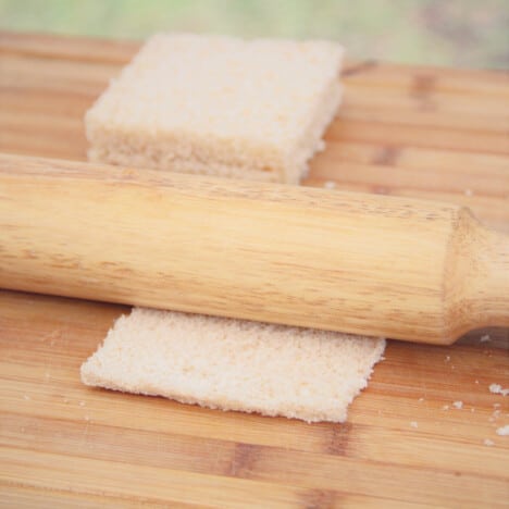 A crust-less slice of whole wheat bread being flattened with a rolling pin on a chopping board.