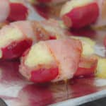 Close up of two fully cooked frankfurters, each oozing melted cheese and wrapped in bacon secured by a toothpick.
