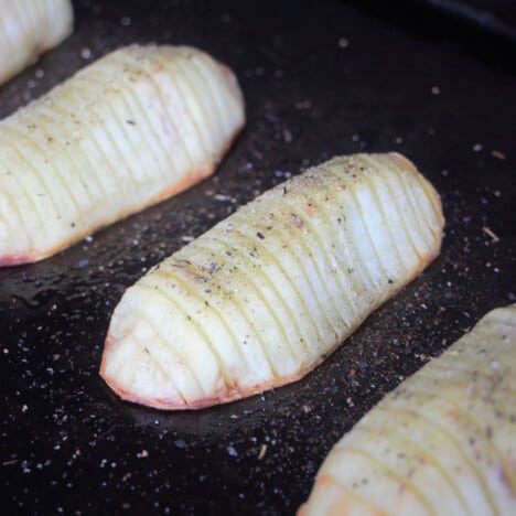 Hasselback potatoes partially cooked on a dark baking tray.