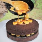 A skillet filled with banana fosters is being tipped over a layer of the chocolate cake.