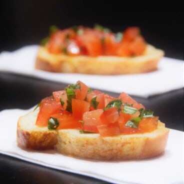 A slice of baguette bread topped with diced tomato and fresh basil on a white napkin, a second bruschetta in the background.