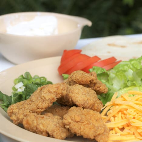 Chicken nuggets, grated cheese, sliced tomatoes, and lettuce laid out on a plate next to tortillas.