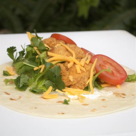 A tortilla topped with cream cheese, chicken nuggets, lettuce, tomato, and cilantro.