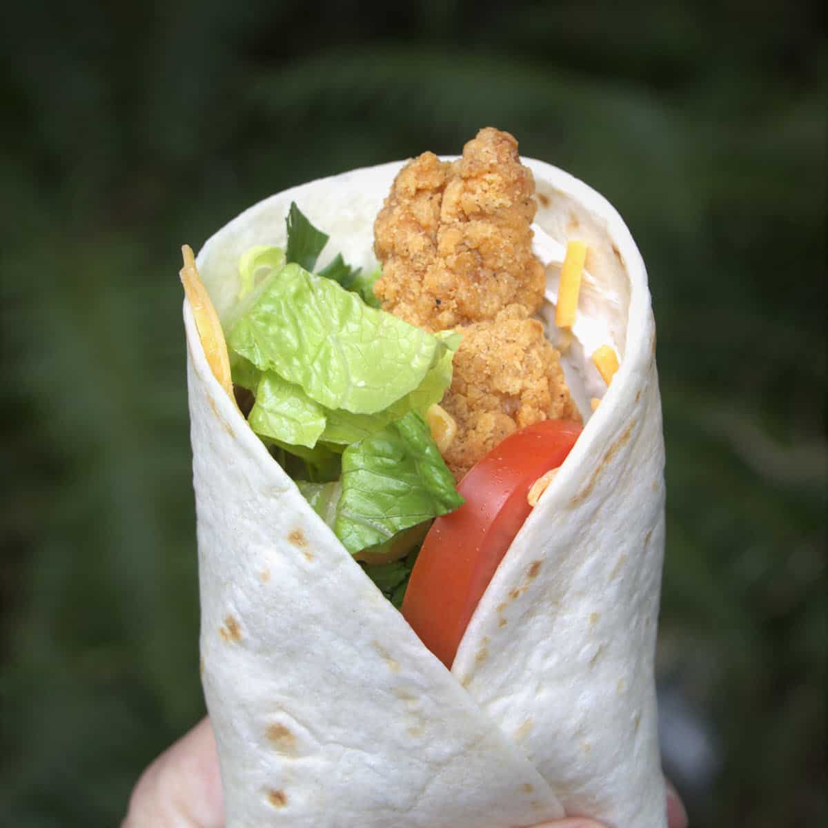 A wrap sliced to reveal chicken nuggets, shredded cheese, red bell pepper and cream cheese rolled up in a wheat tortilla.