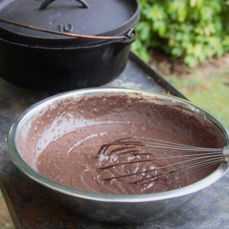 A stainless bowl with the chocolate cake batter in it with a whisk sitting in front of a Dutch oven.